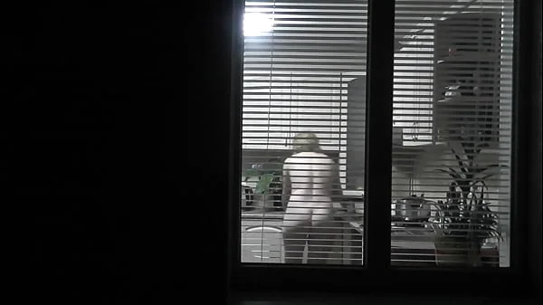 Peeping. Voyeur. Neighbor pervert voyeur in evening on street looks out window as naked sexy neighbor in kitchen prepares dinner for her husband. Naked in public. Naked at home. Family.  Outdoor